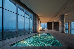 Una piscina interior moderna con suelo de baldosas, ventanas amplias que ofrecen vistas a la ciudad y un área de descanso con asientos.
