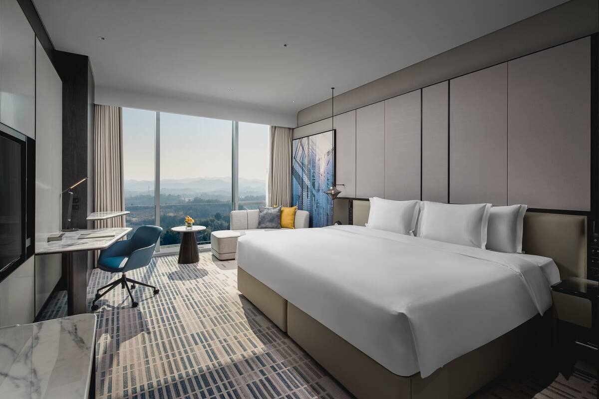 A modern hotel room with a large bed, a desk, a chair, and a view of the cityscape.