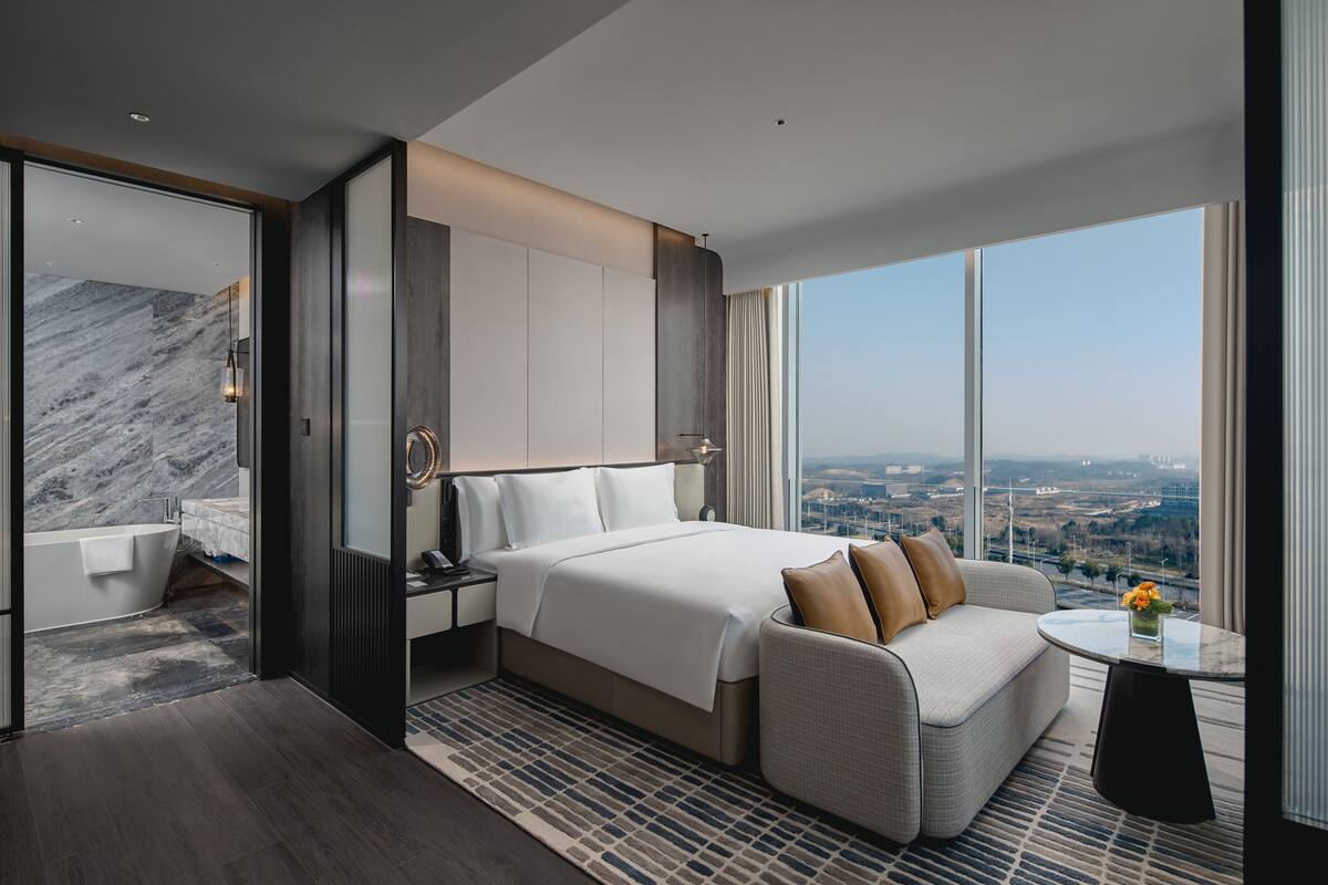 A modern hotel room with a large bed, a sofa, a small table with flowers, and a city view.