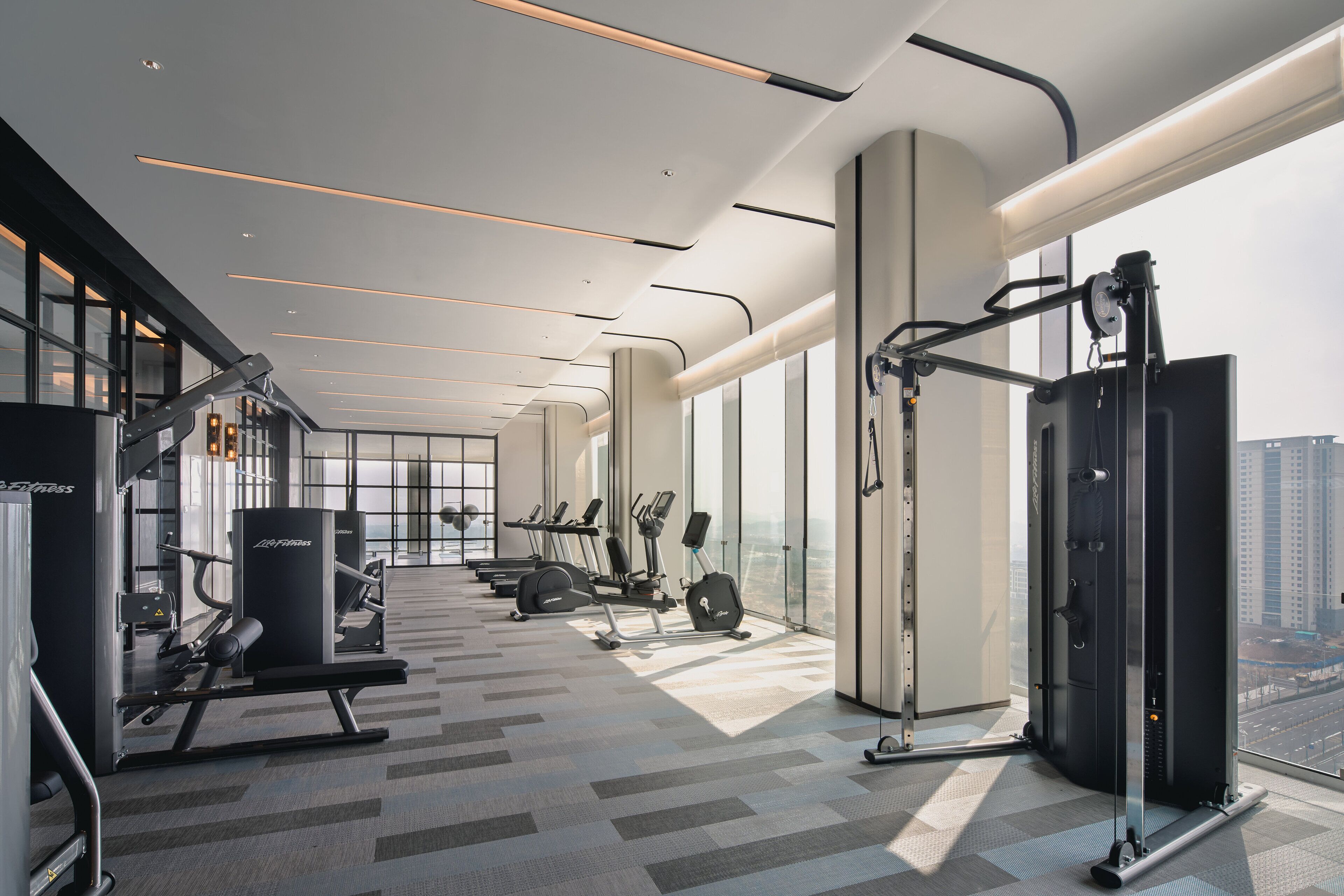 Ett modernt gym med olika typer av träningsutrustning, stora fönster och utsikt över staden.