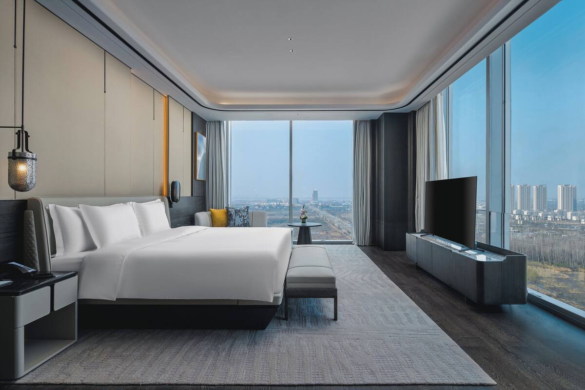 A modern hotel room with a large bed, a flat-screen TV, a small table, and a view of the city.