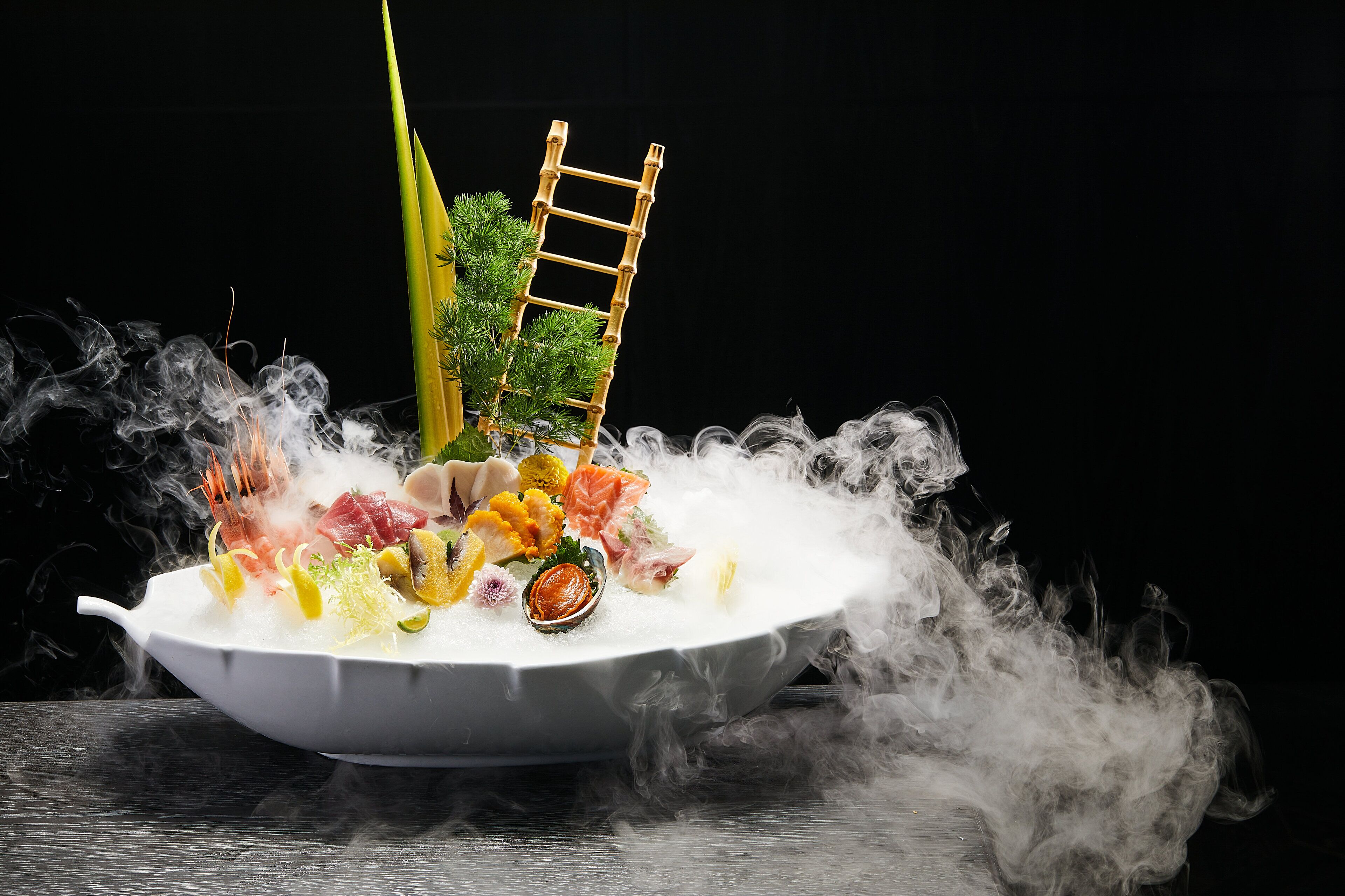 A white ceramic dish filled with various seafood and vegetables, surrounded by smoke, with a bamboo ladder and greenery on top.
