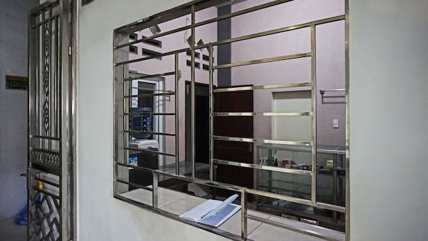 A room with a window featuring a metal grid design, a visible door with a decorative pattern, and a shelf with various items.