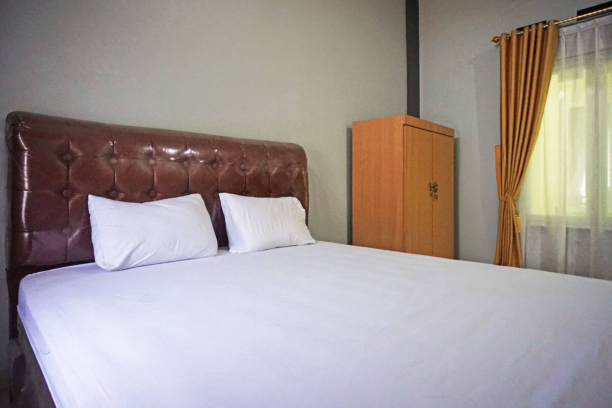 A neatly made bed with a brown headboard and white bedding in a room with a wooden wardrobe and a window with curtains.