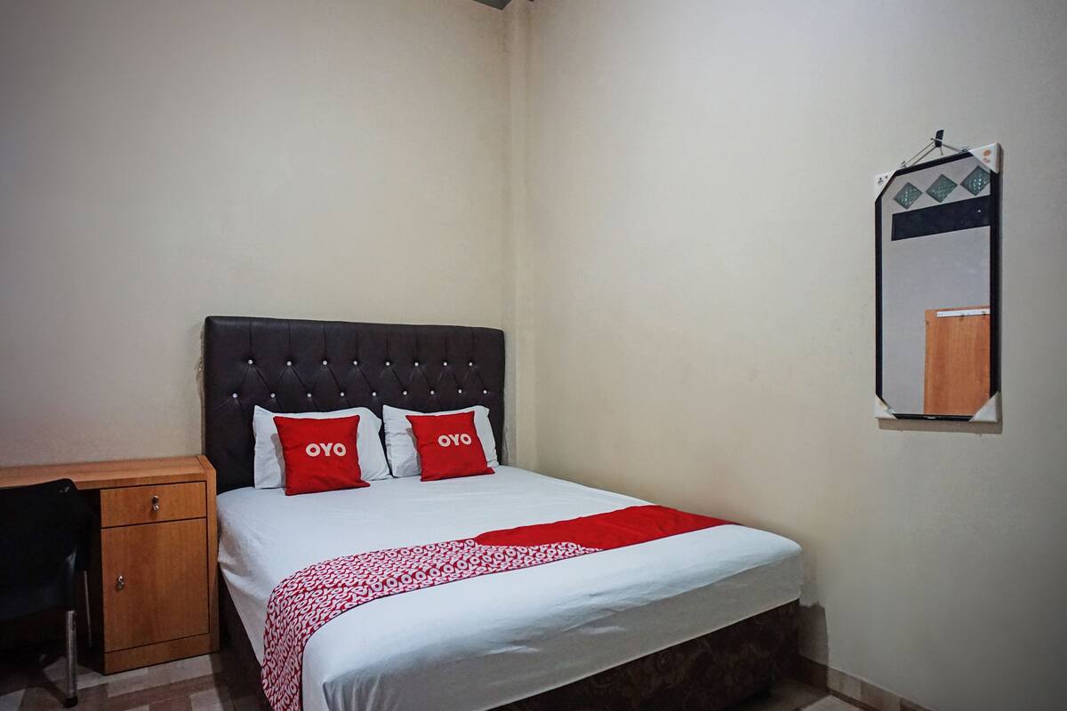 A neatly made bed with a patterned bedspread and two red pillows, a wooden nightstand, and a mirror on the wall.