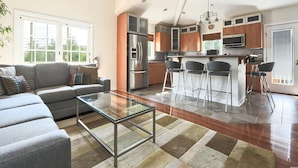 A modern living room with a glass coffee table, a grey sofa, and a kitchen with a stainless steel refrigerator and wooden cabinets. - 6 BR, Pool, Hot Tub and 5 min to Ocean (Loveladies)