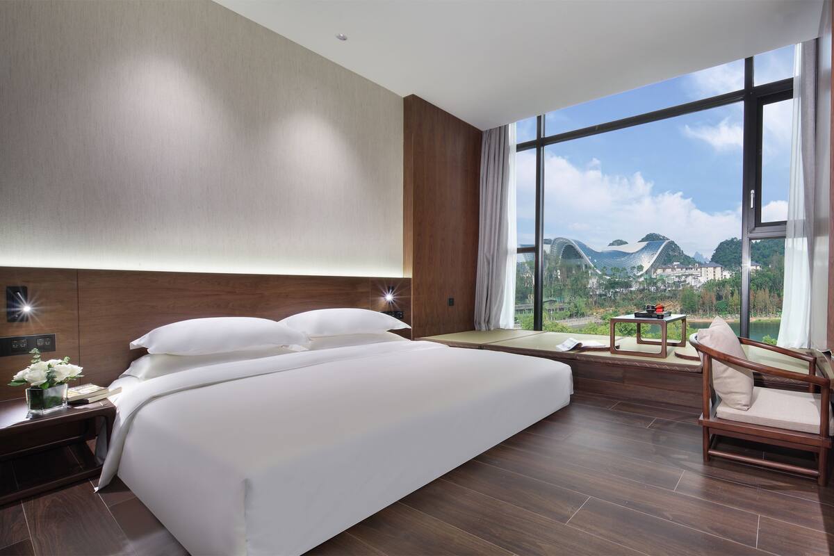 A hotel room with a large bed, a wooden headboard, a view of mountains, and a small table with a chair.