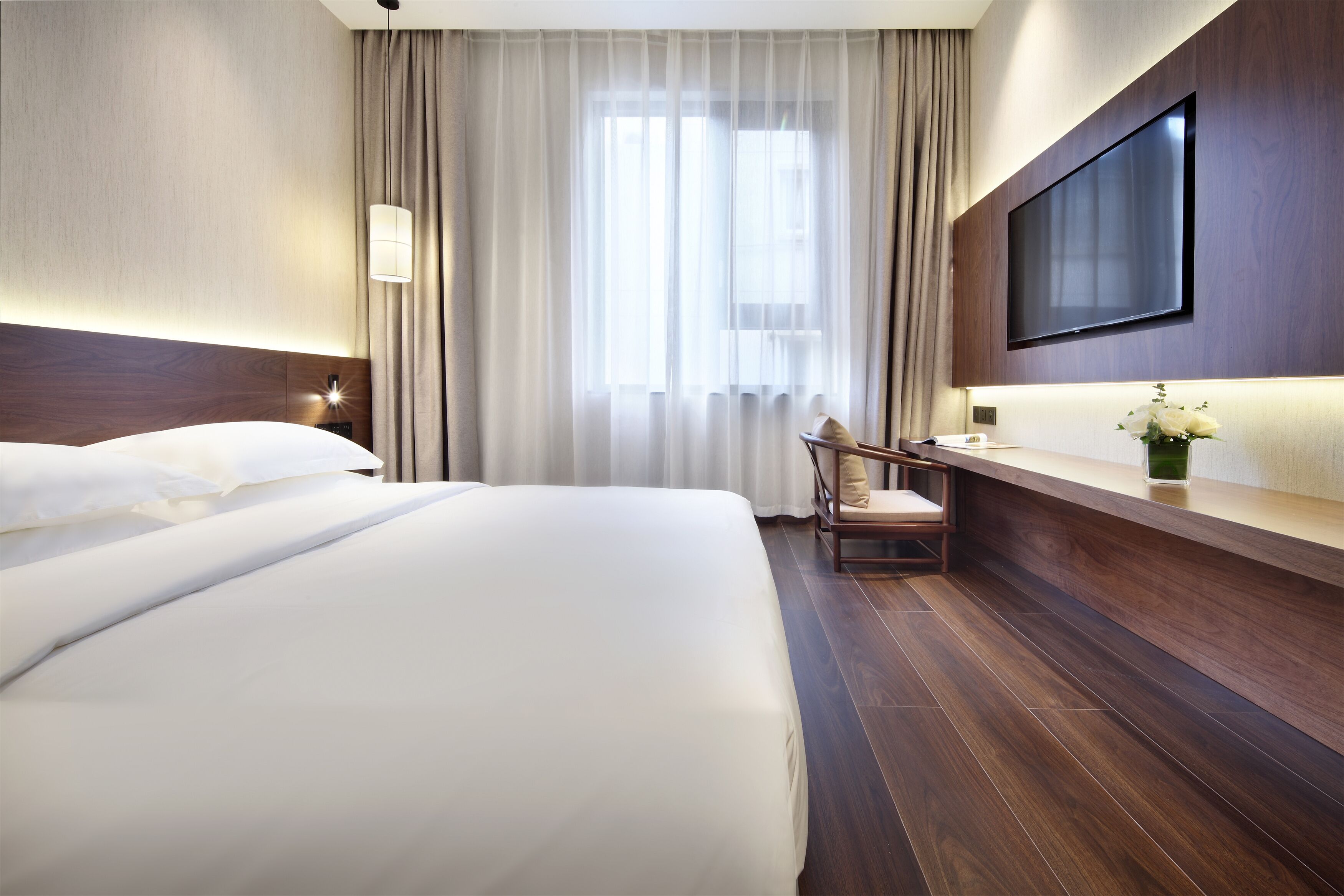 A hotel room with a large bed, a desk with a chair, a flat-screen TV mounted on the wall, and a window with curtains.