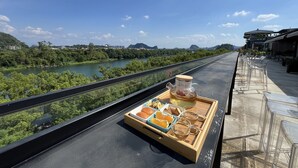 Eine Terrasse mit einem hölzernen Tablett, das Teekanne, Tassen und Snacks präsentiert, mit Blick auf einen Fluss und grüne Vegetation.