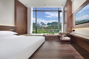 A modern hotel room with a large bed, a desk, and a view of a mountainous landscape.