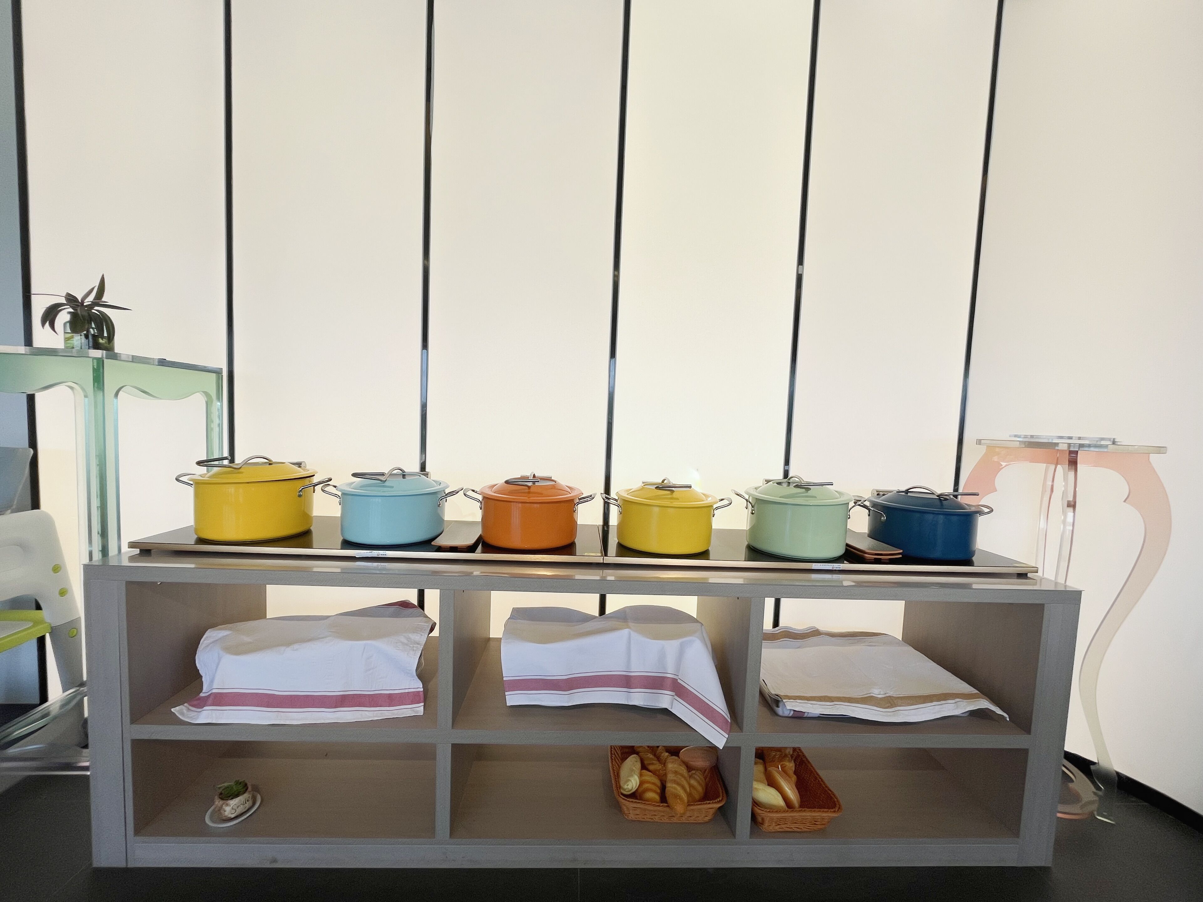 Una encimera de cocina con seis ollas de colores y dos estantes inferiores con manteles doblados y cestas de pan.