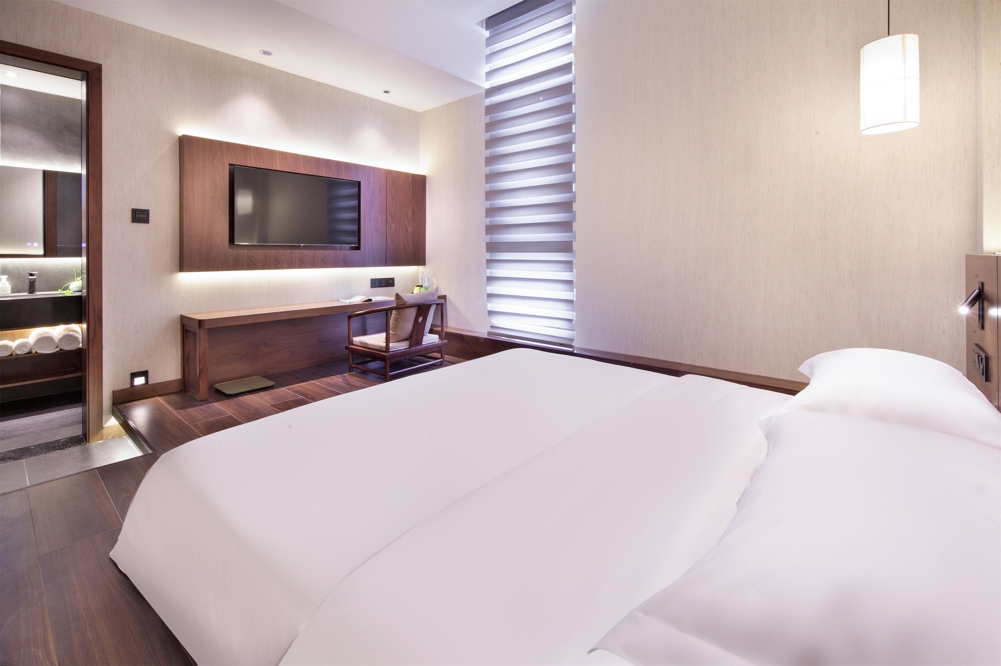 A modern hotel room with a large bed, a desk with a chair, a flat-screen TV mounted on the wall, and a window with blinds.