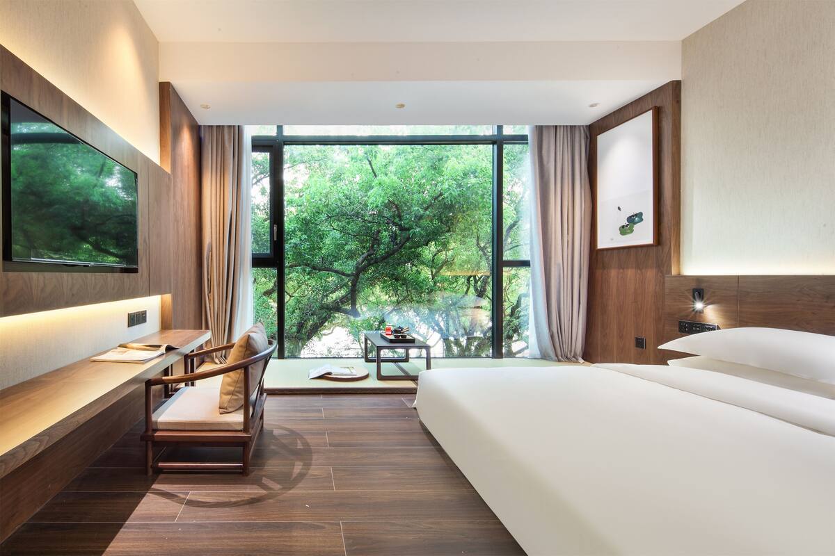 A modern hotel room with a large bed, a desk with a chair, a TV mounted on the wall, and a large window offering a view of trees.