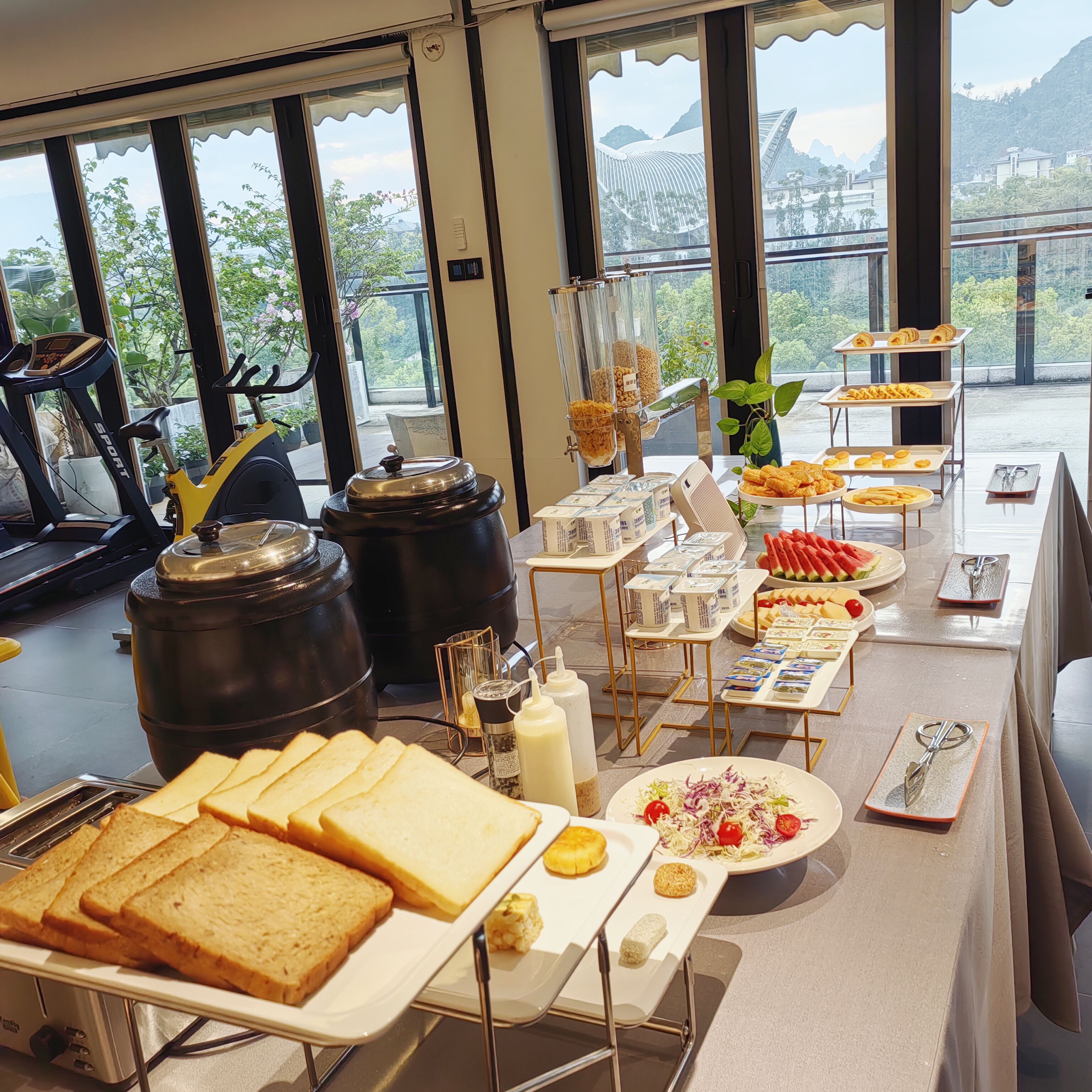 Un bufé de desayuno con diversas opciones de comida, una cinta de correr y una vista panorámica a través de amplios ventanales.