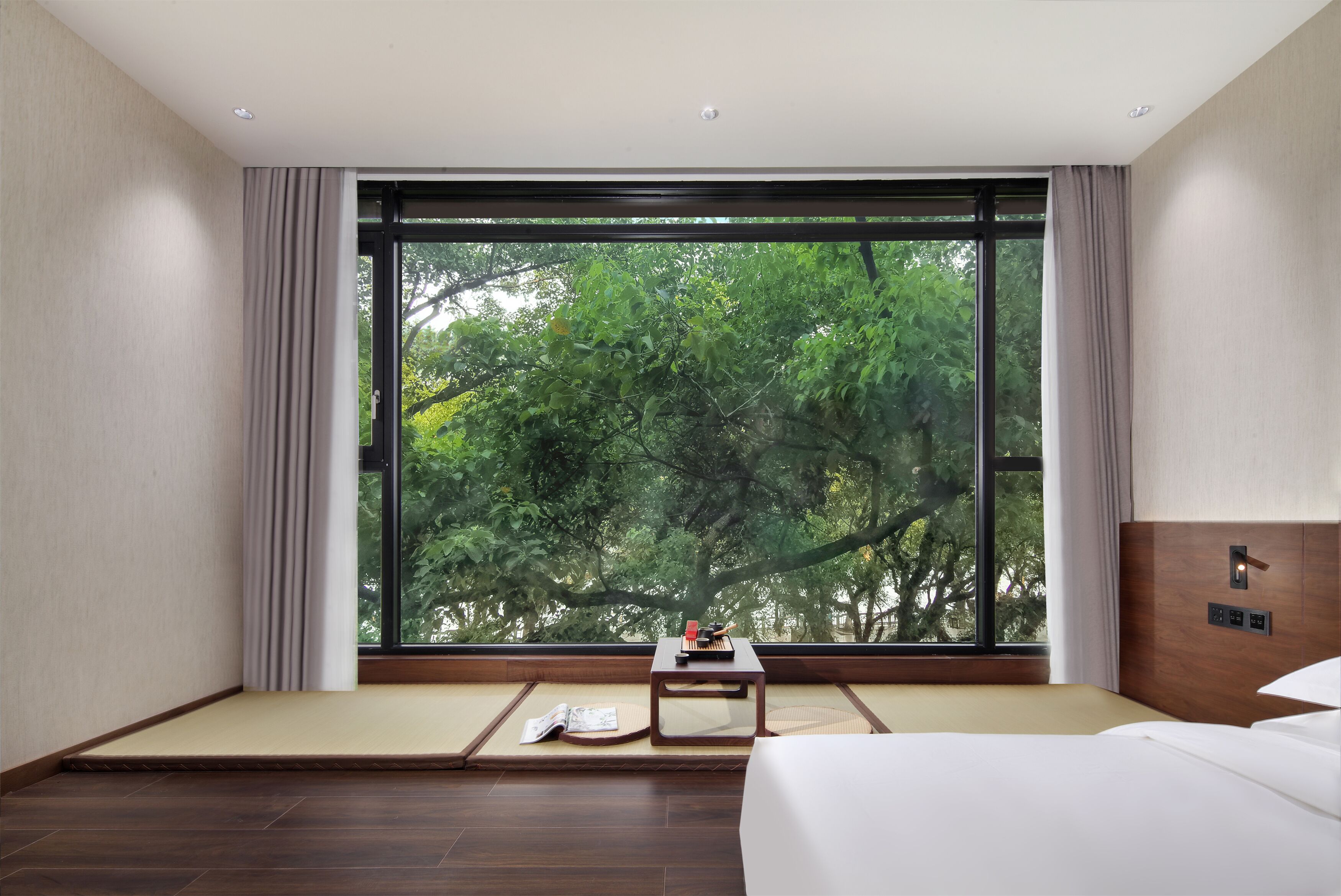 A modern hotel room with a large window, a bed, a small table with a magazine, and a view of trees outside.
