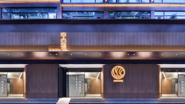 Ein moderner Hotelaufgang mit auffälligem Logo, großen Holzpaneelen und einer gut beleuchteten Fassade.