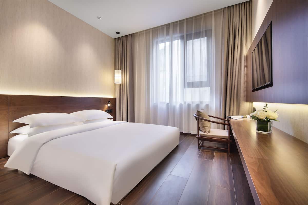 A hotel room with a large bed, a desk with a chair, a vase of flowers, and a large window with curtains.