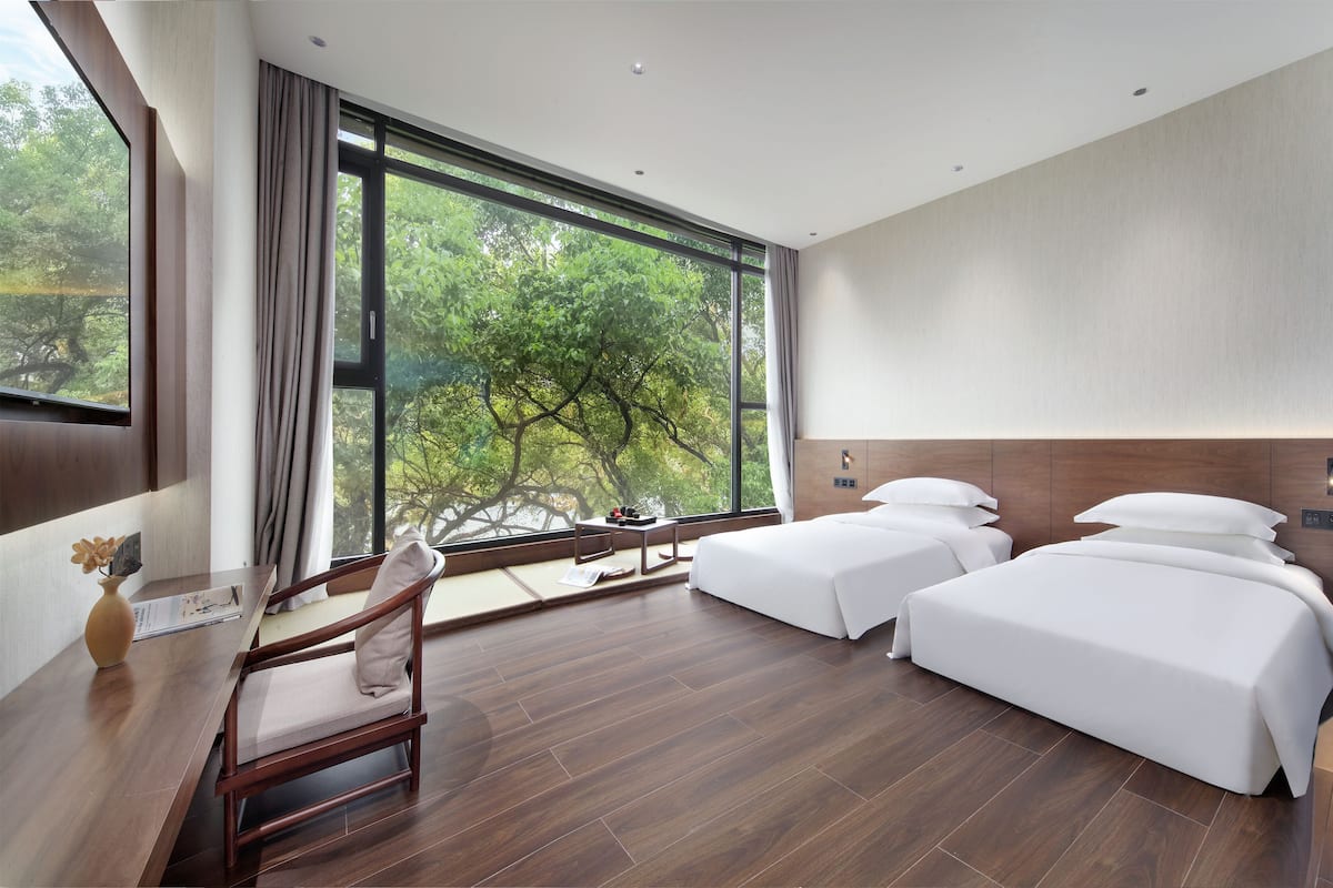 A modern hotel room with two single beds, a wooden desk, a chair, and a large window offering a view of trees.