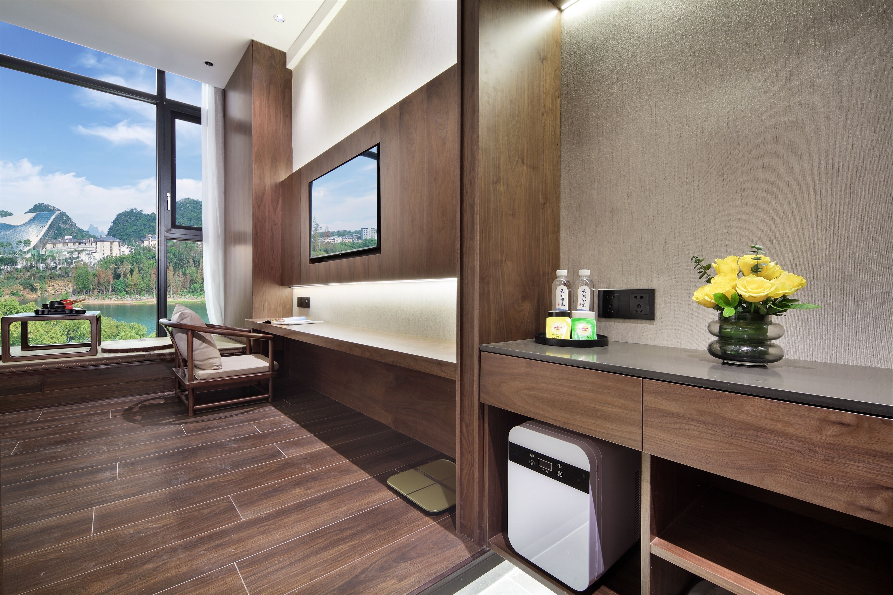 Una habitación moderna con un gran ventanal, un televisor de pantalla plana, un escritorio de madera, una silla, una mesita y un jarrón con flores amarillas.