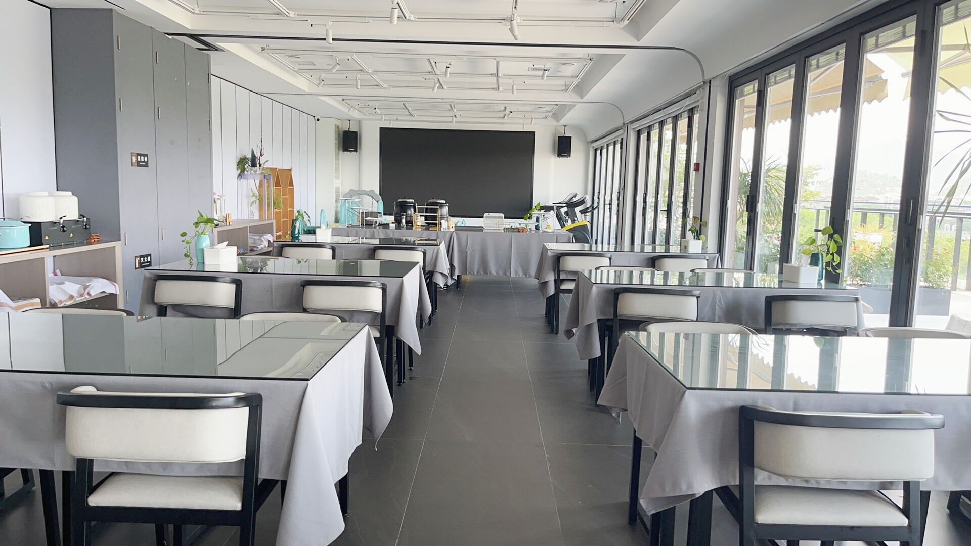 A modern restaurant with long tables and chairs, large windows, and a stage at the front.