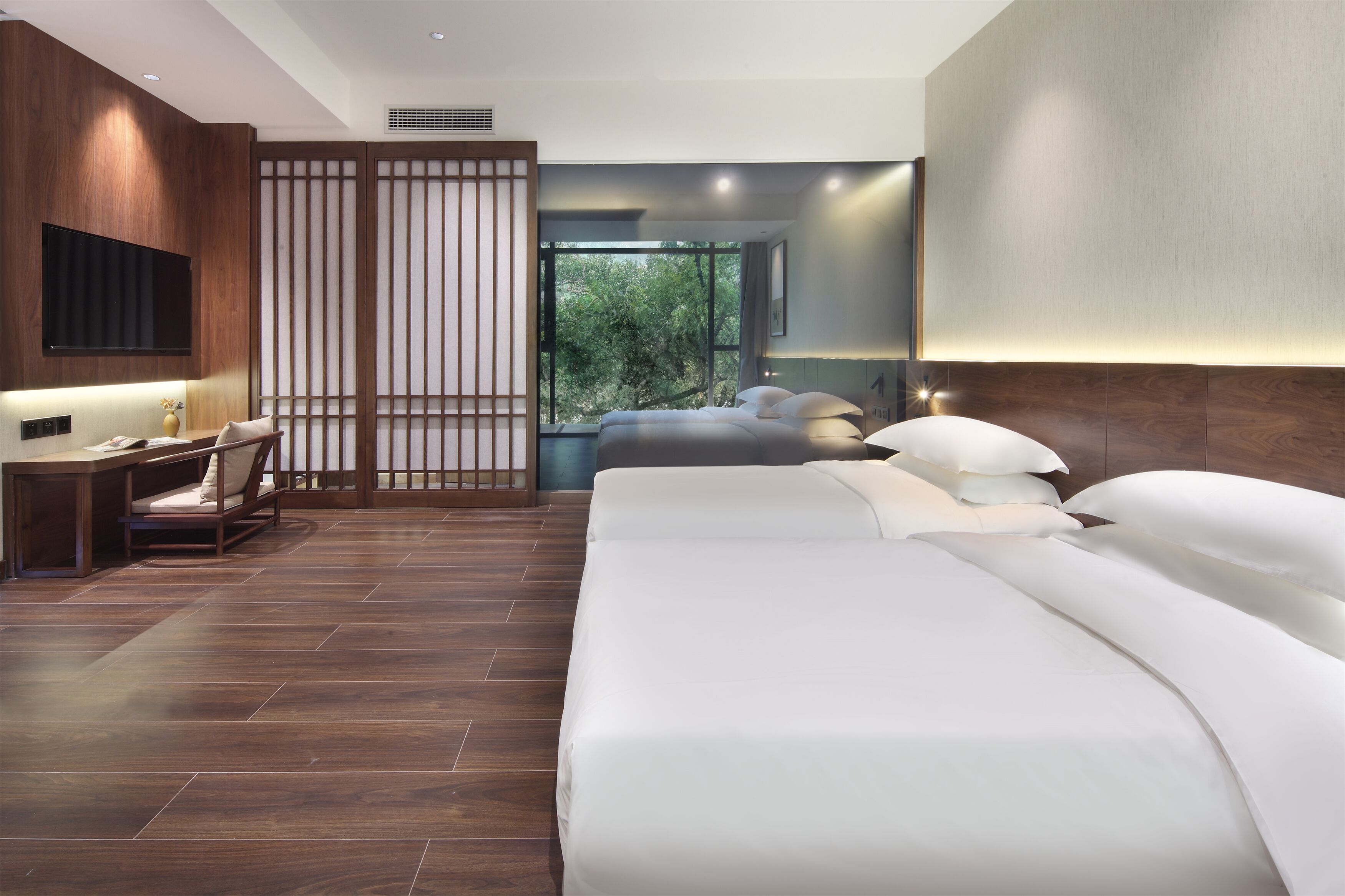 Een moderne hotelkamer met een groot bed, een bureau met stoel, een televisie en een groot raam met uitzicht op bomen.