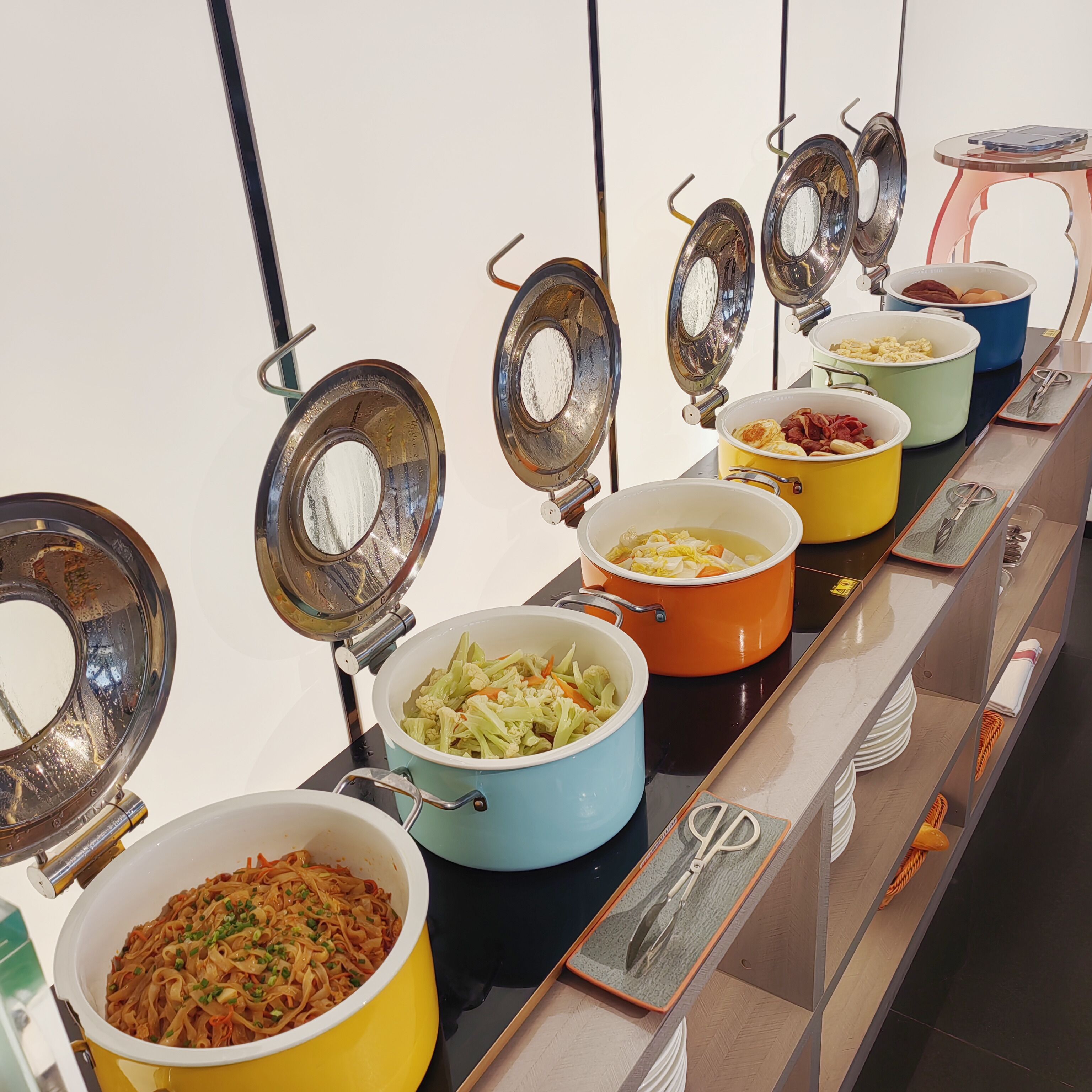 A buffet line with various dishes in colorful pots, including pasta, vegetables, and eggs.