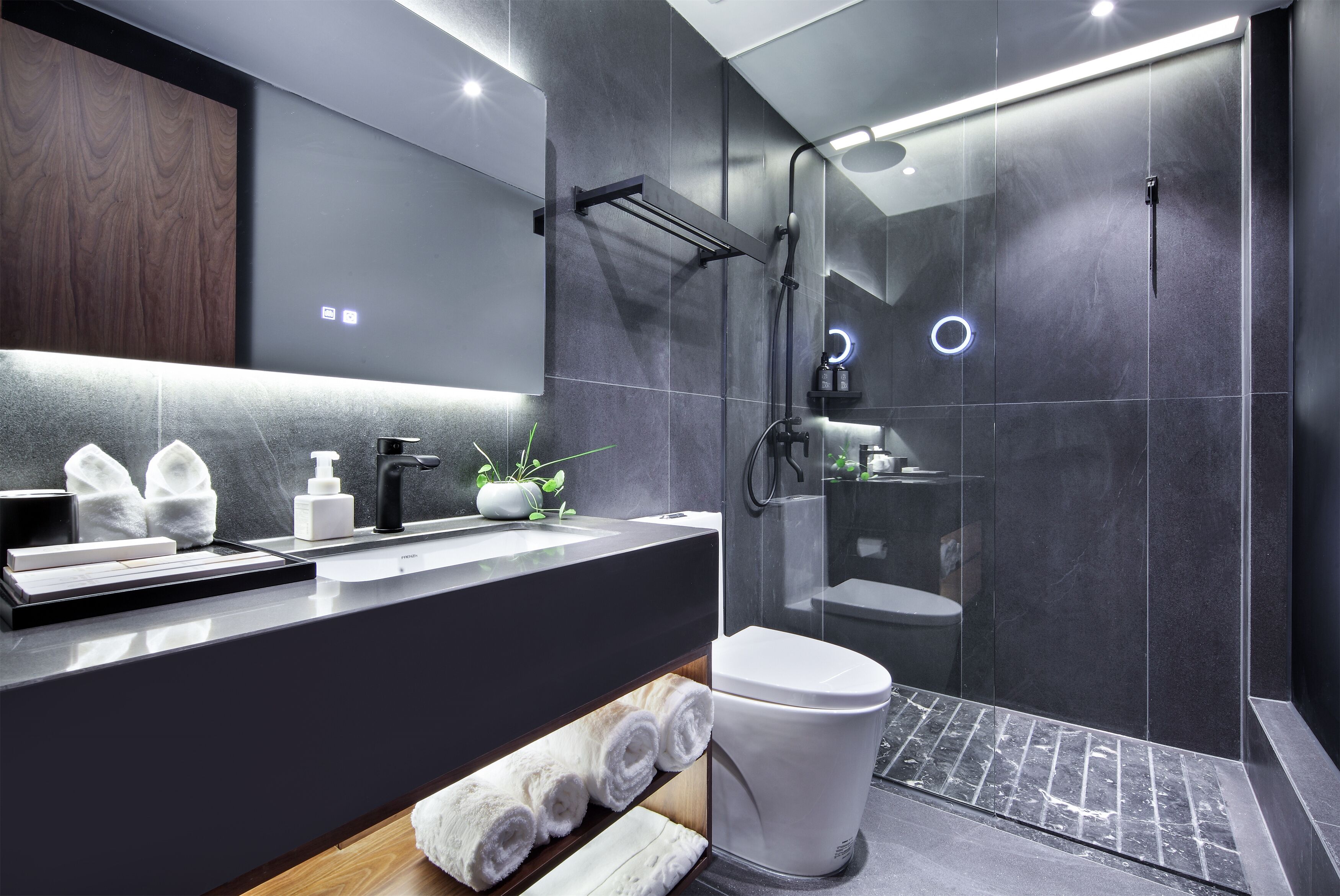 A modern bathroom with a dark tile wall, a wooden vanity, a white toilet, and a glass-enclosed shower area.