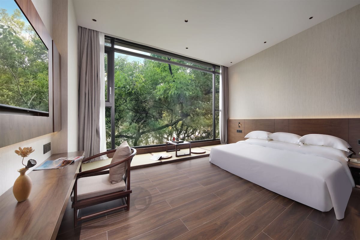 A modern hotel room with a large bed, a desk with a chair, and a view of trees through large windows.