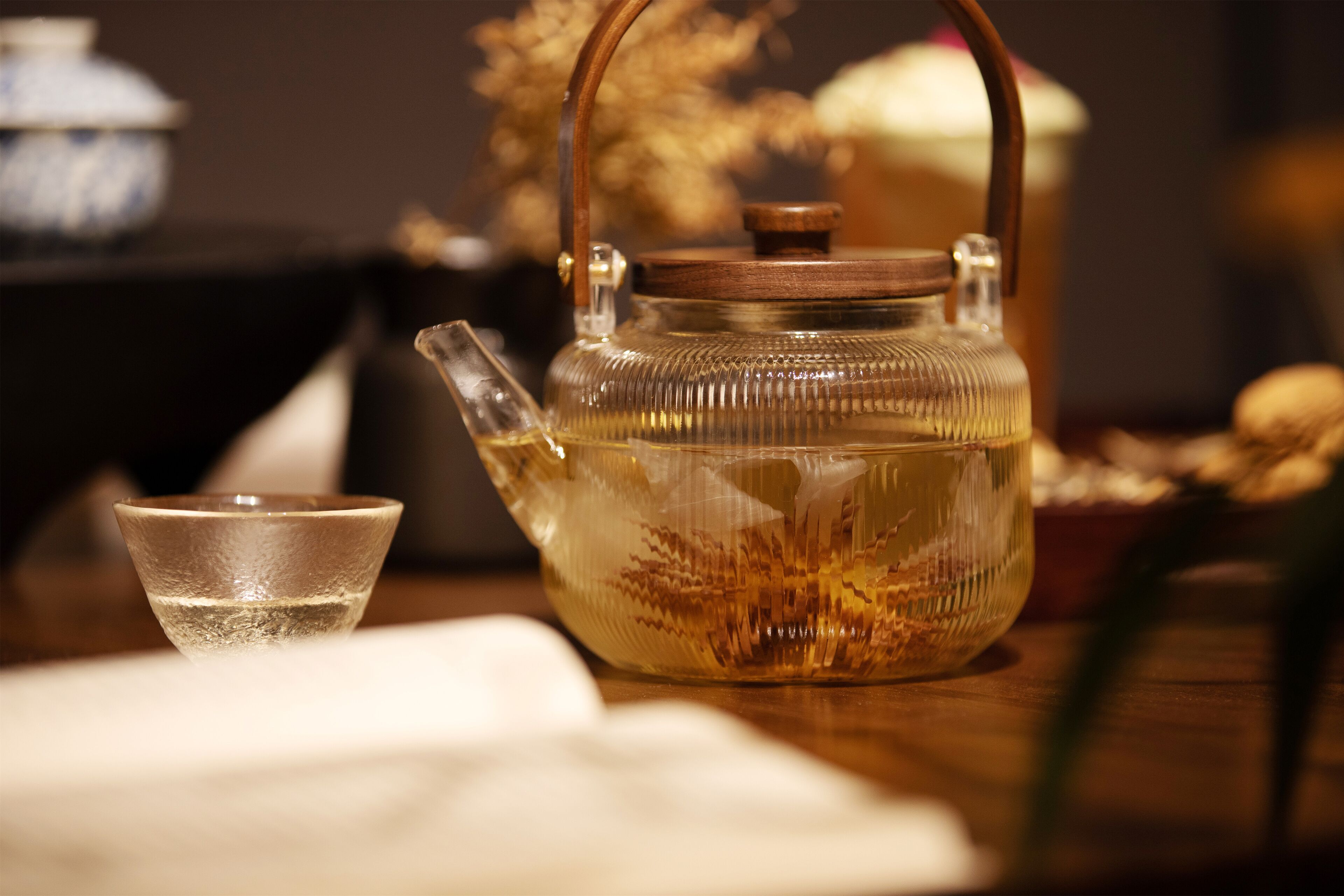 Une théière en verre transparent avec un couvercle et une anse en bois, une petite tasse en verre et une table en bois sur un fond flou.