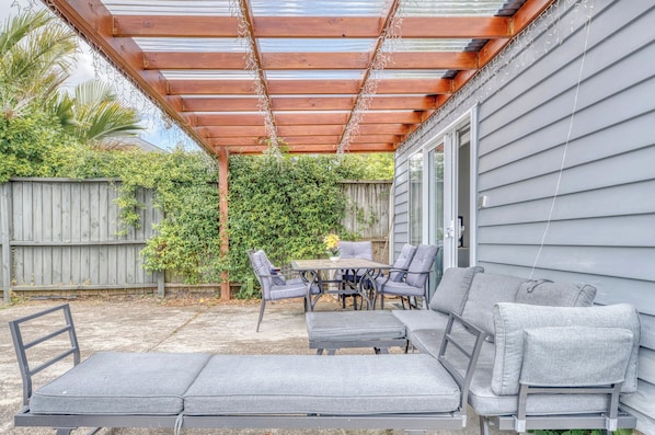 En overdækket terrasse med et bord, stole og en bænk, samt en træpergola.