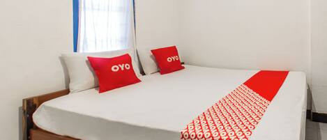 Una cama con ropa blanca y almohadas rojas, cabecera de madera y una ventana con cortinas azules.