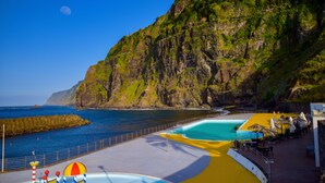 A coastal area with a rocky cliff, a swimming pool, and a beach area with umbrellas and chairs. - Paradise town with ocean pool, mountains, waterfalls, surfing, shops and bars.  (Ponta Delgada svc 2879-S Vicente)