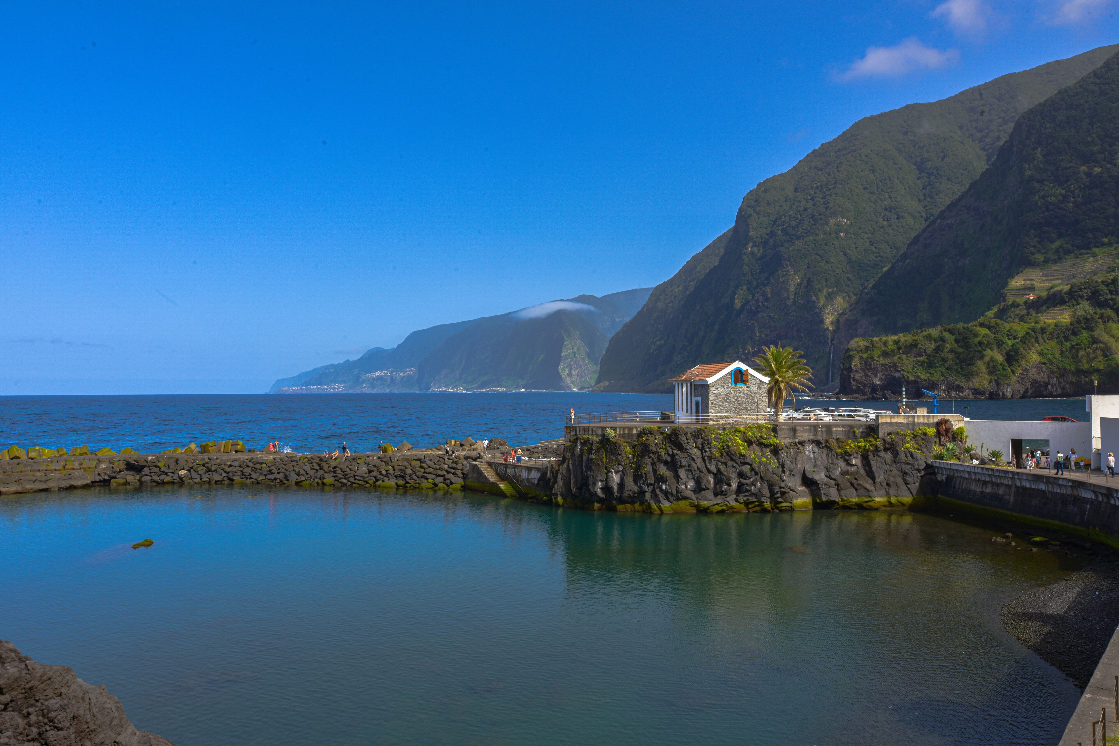 A coastal scene with a small building on a rocky outcrop, calm water, and mountains in the background.