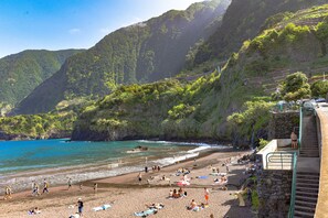 A beach with people sunbathing and swimming, surrounded by lush green hills and a clear blue sea.