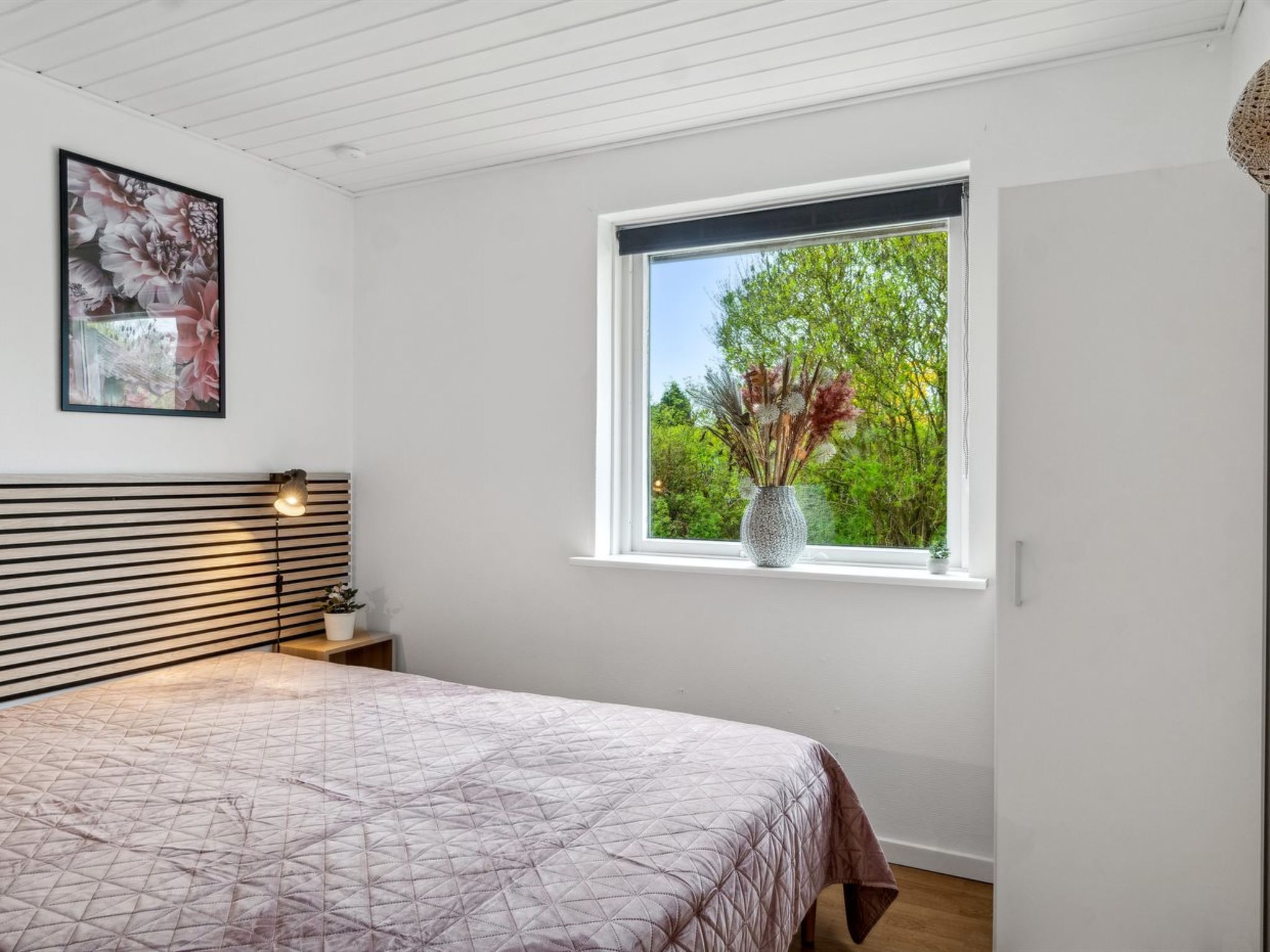 Une chambre à coucher avec un lit, une fenêtre donnant sur un paysage verdoyant, un tableau accroché au mur et une lampe de chevet.