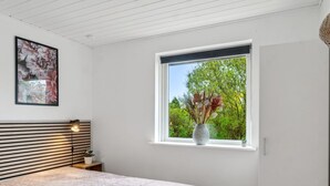 A bedroom with a bed, a window with a view of greenery, a wall-mounted picture, and a bedside lamp.
