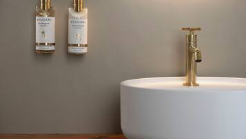 Une salle de bain avec un lavabo blanc et un robinet doré, deux flacons de lotion pour le corps BVLGARI et une table en bois.