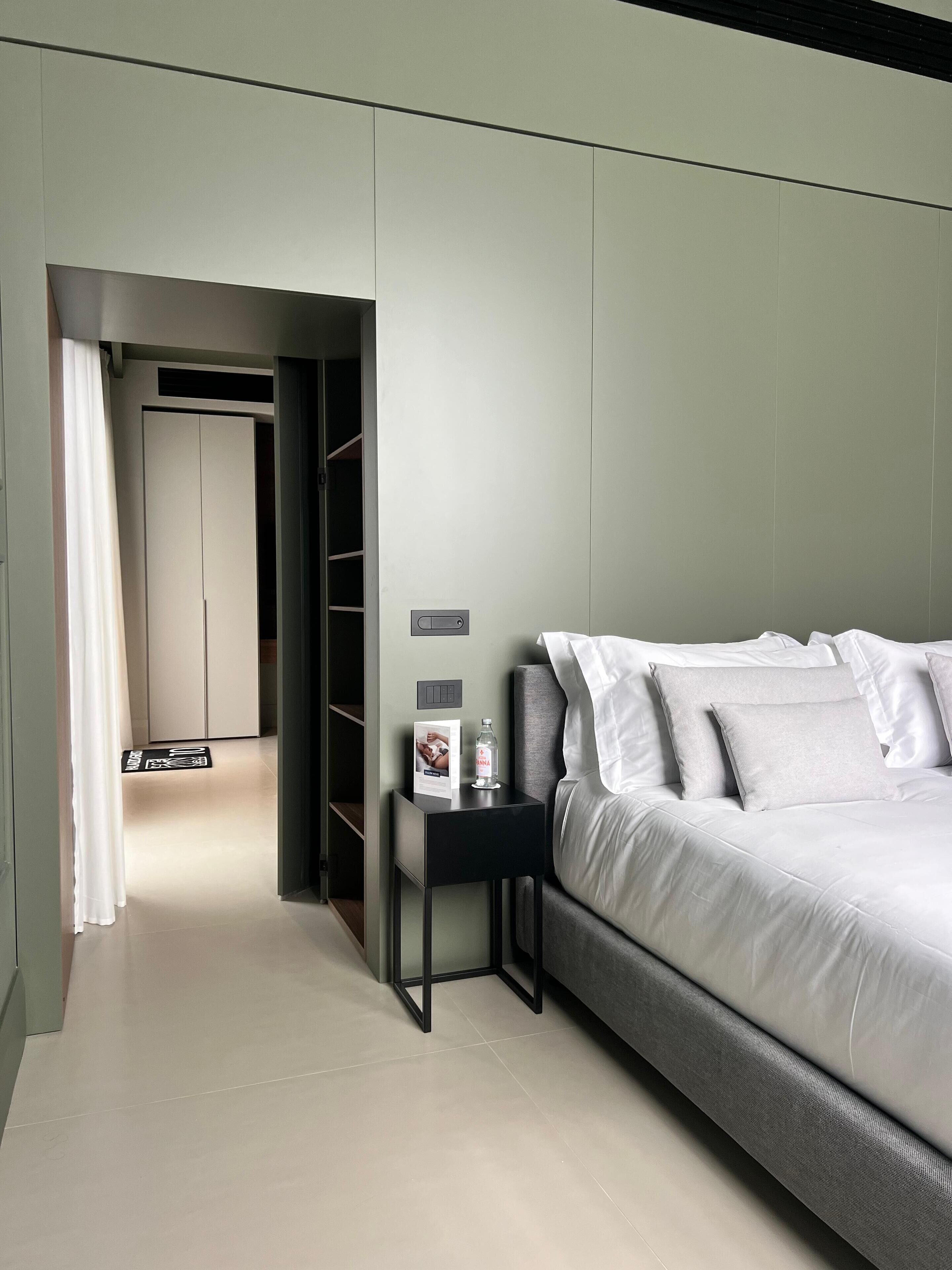 A modern hotel room with a grey sofa, a small bedside table with a bottle and a card, and a wardrobe in the background.
