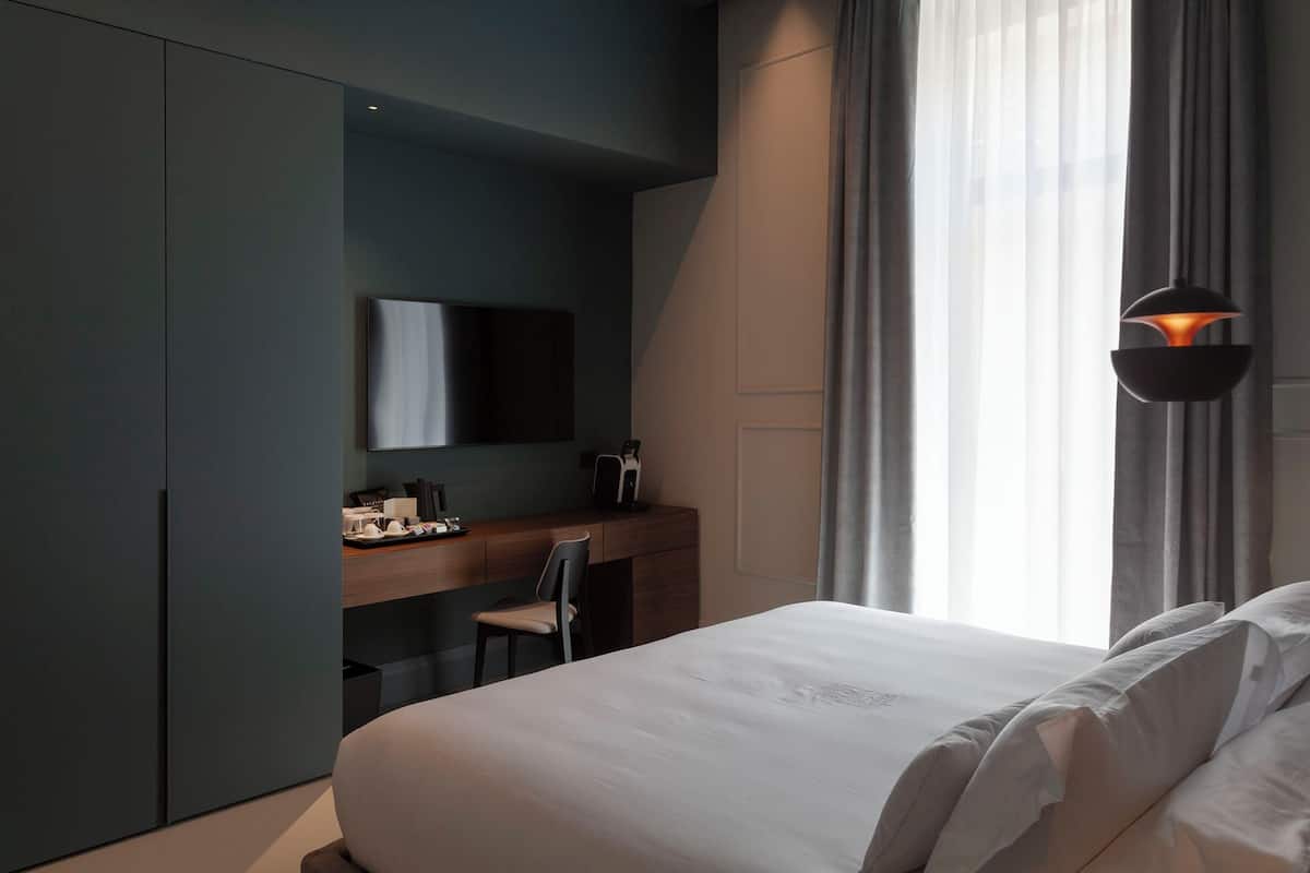 Una habitación de hotel moderna con una cama, un escritorio, una silla, un televisor de pantalla plana y un ventanal con cortinas.