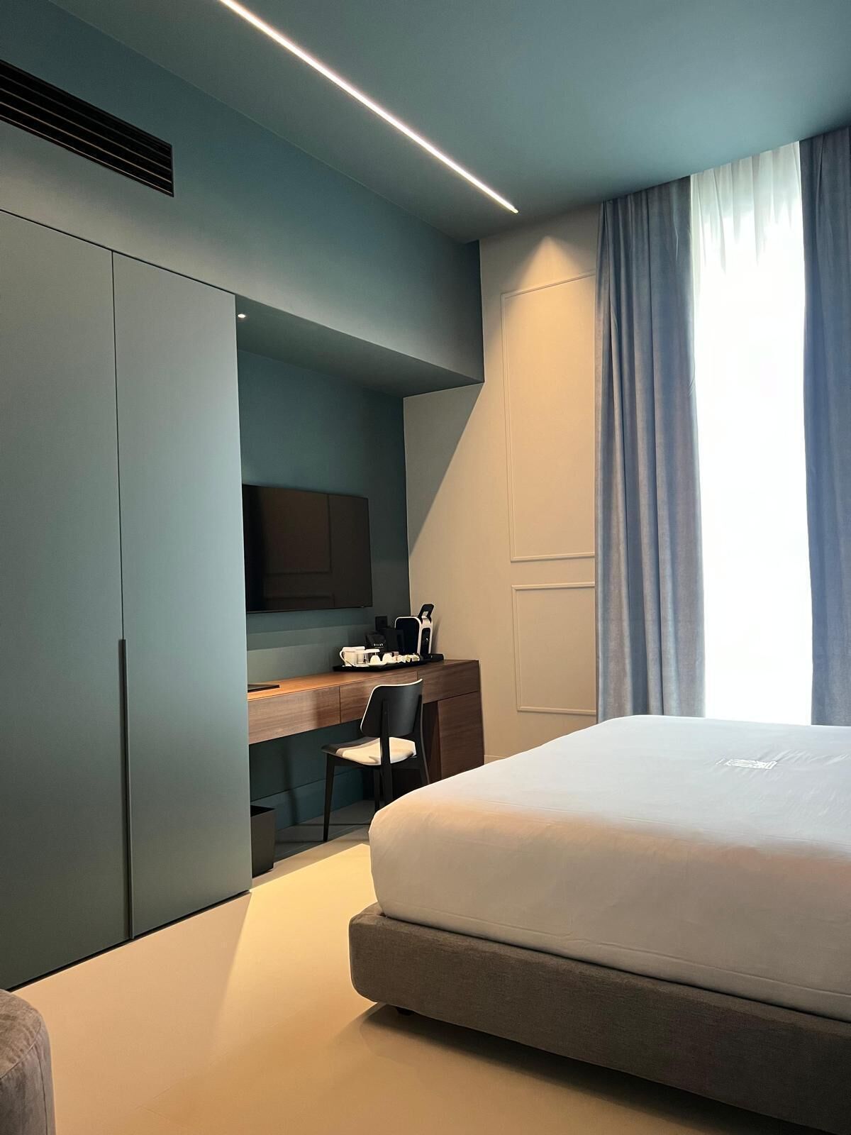 A modern hotel room with a large bed, a desk with a chair, and a wardrobe.