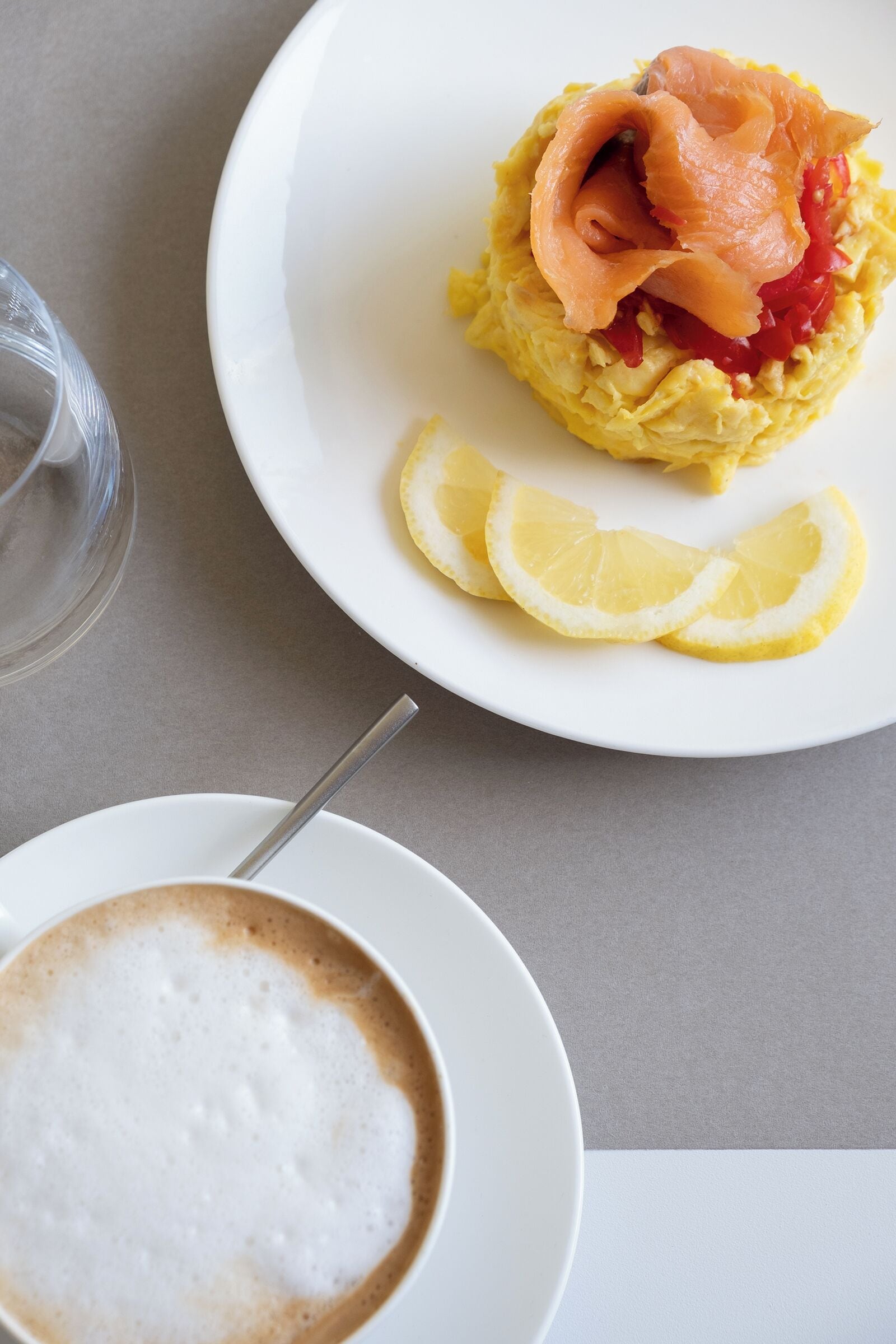 A breakfast setting with scrambled eggs topped with smoked salmon and red peppers, accompanied by lemon slices and a cup of coffee.