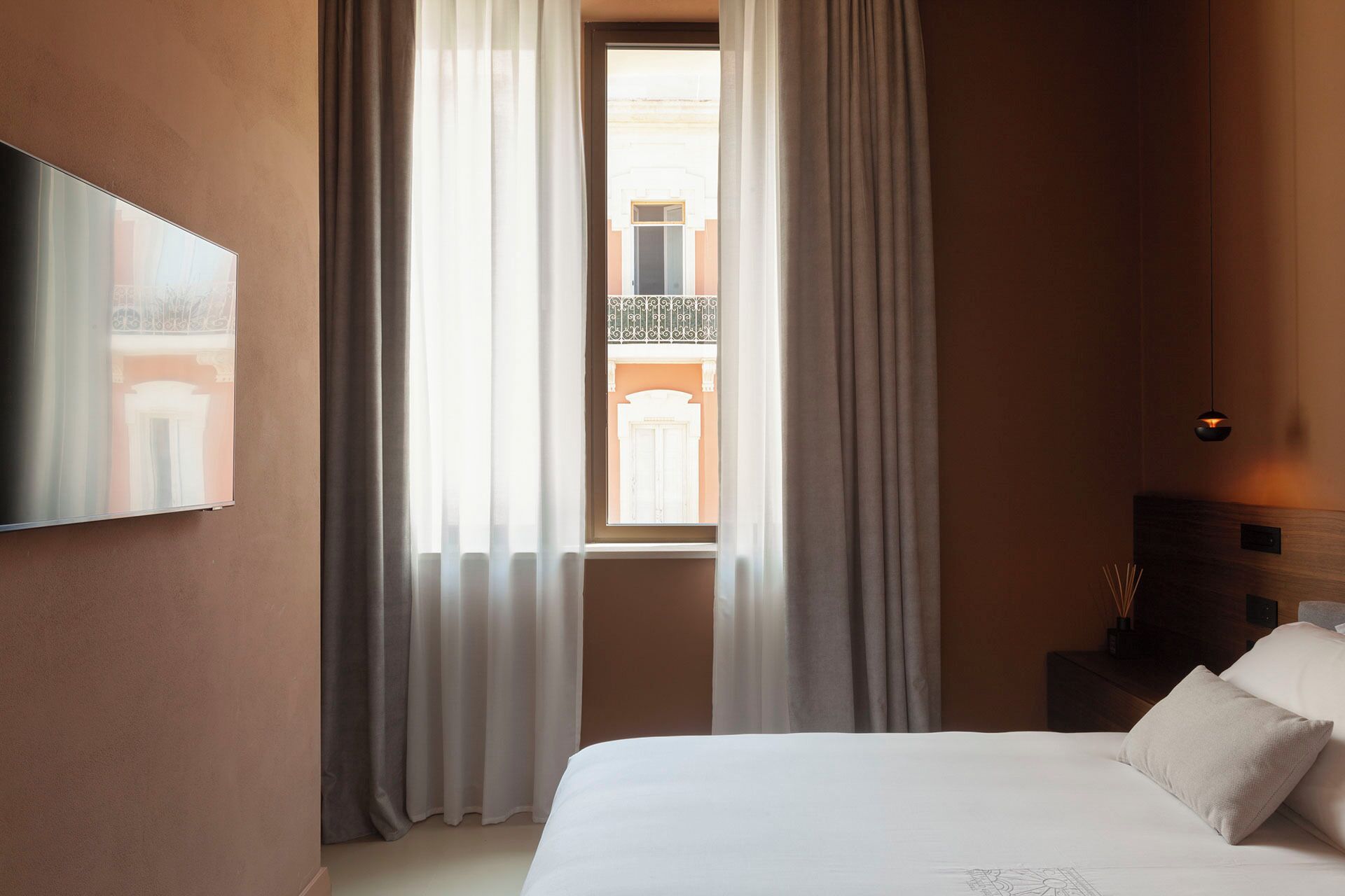A hotel room with a bed, curtains, a painting on the wall, a window with a view of the outside, and a bedside table with a lamp.