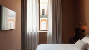 A hotel room with a bed, curtains, a painting on the wall, a window with a view of the outside, and a bedside table with a lamp.