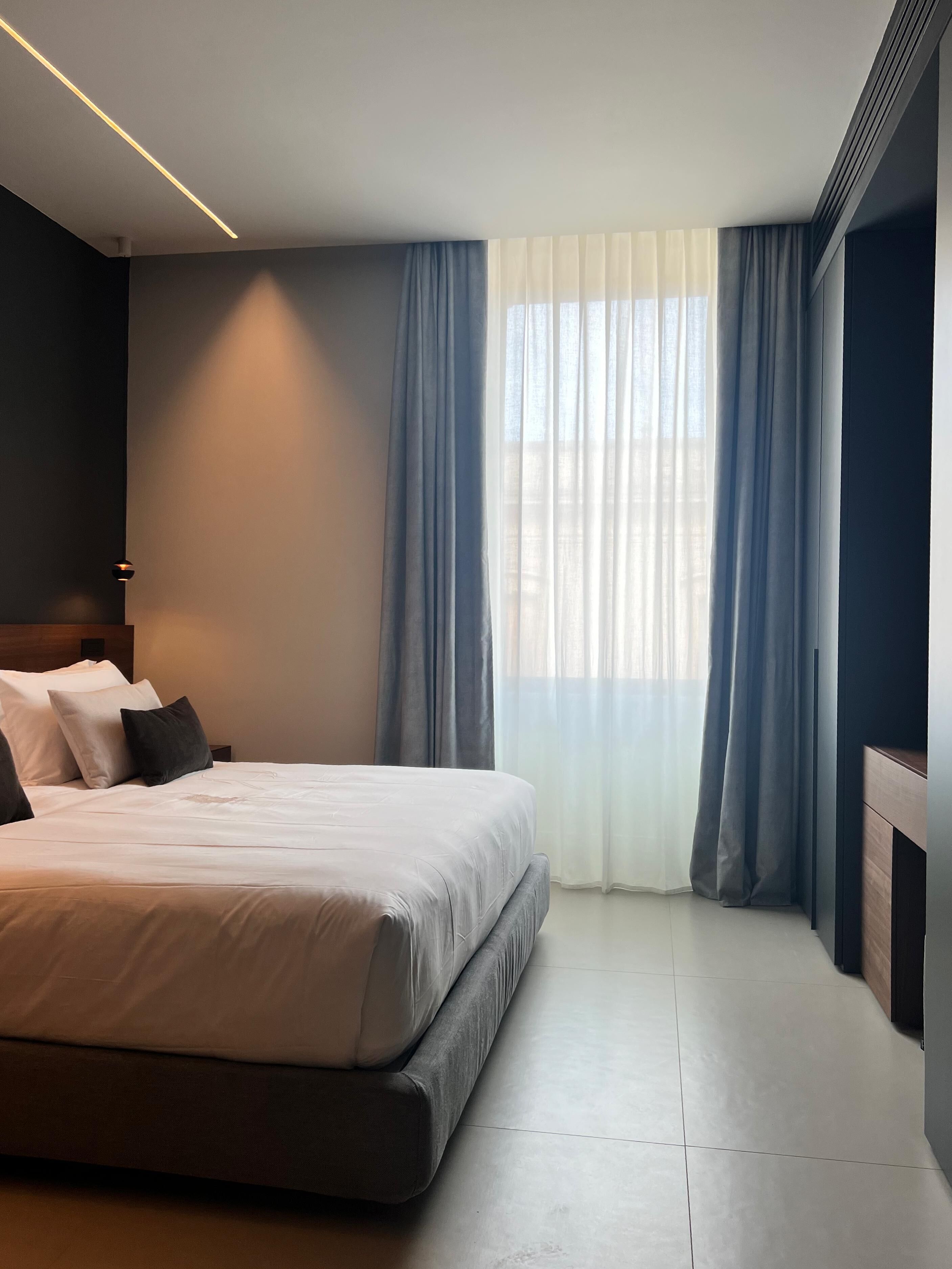A modern hotel room with a large bed, a headboard, and a view of the city through sheer curtains.