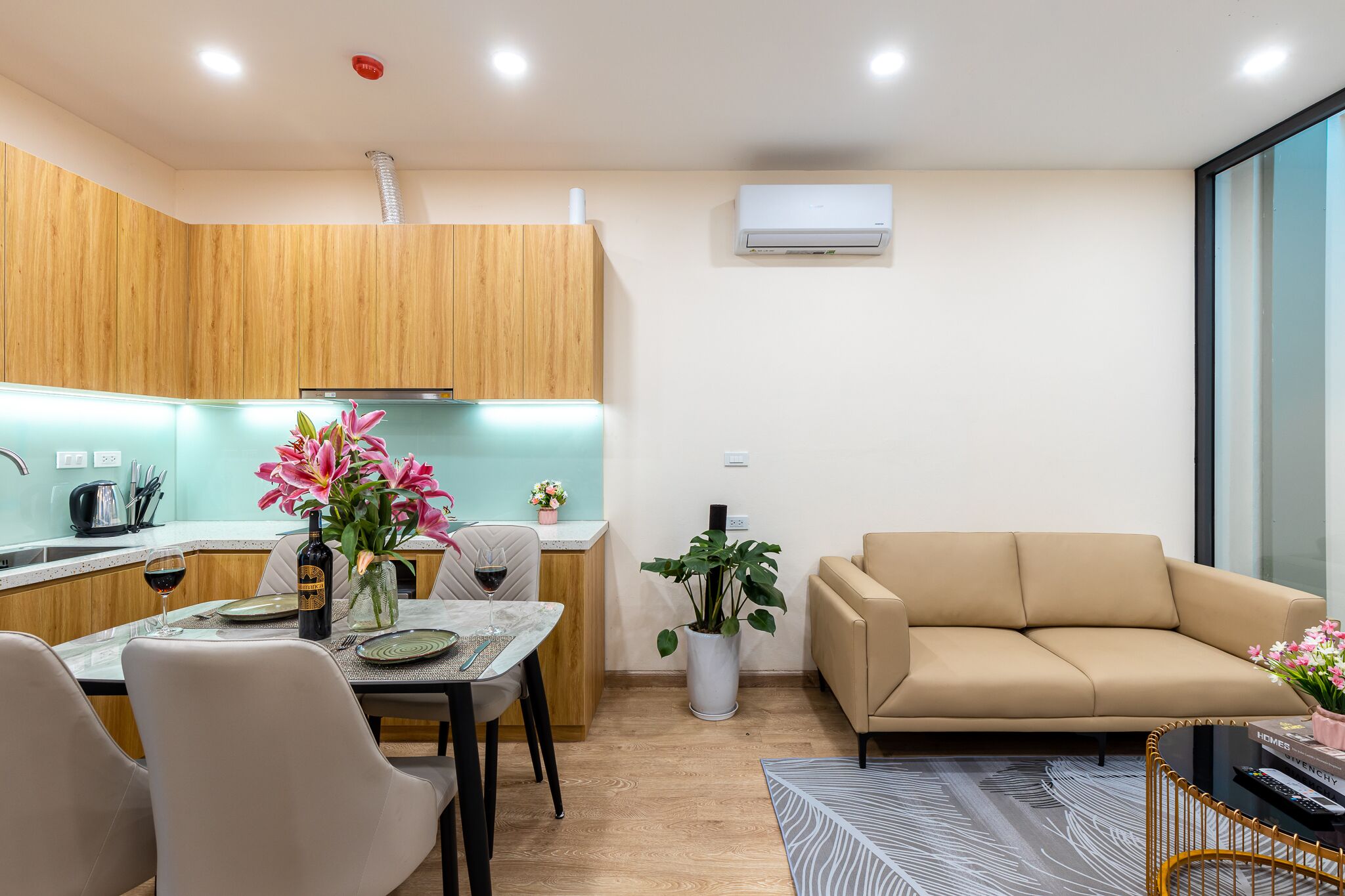 A modern kitchen with wooden cabinets, a dining table with chairs, a sofa, and a potted plant.