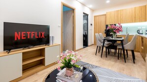 Un saló modern amb televisió de pantalla plana que mostra 'NETFLIX', una zona de menjador amb taula i cadires, i una cuina amb armaris de fusta i un rentapeus.