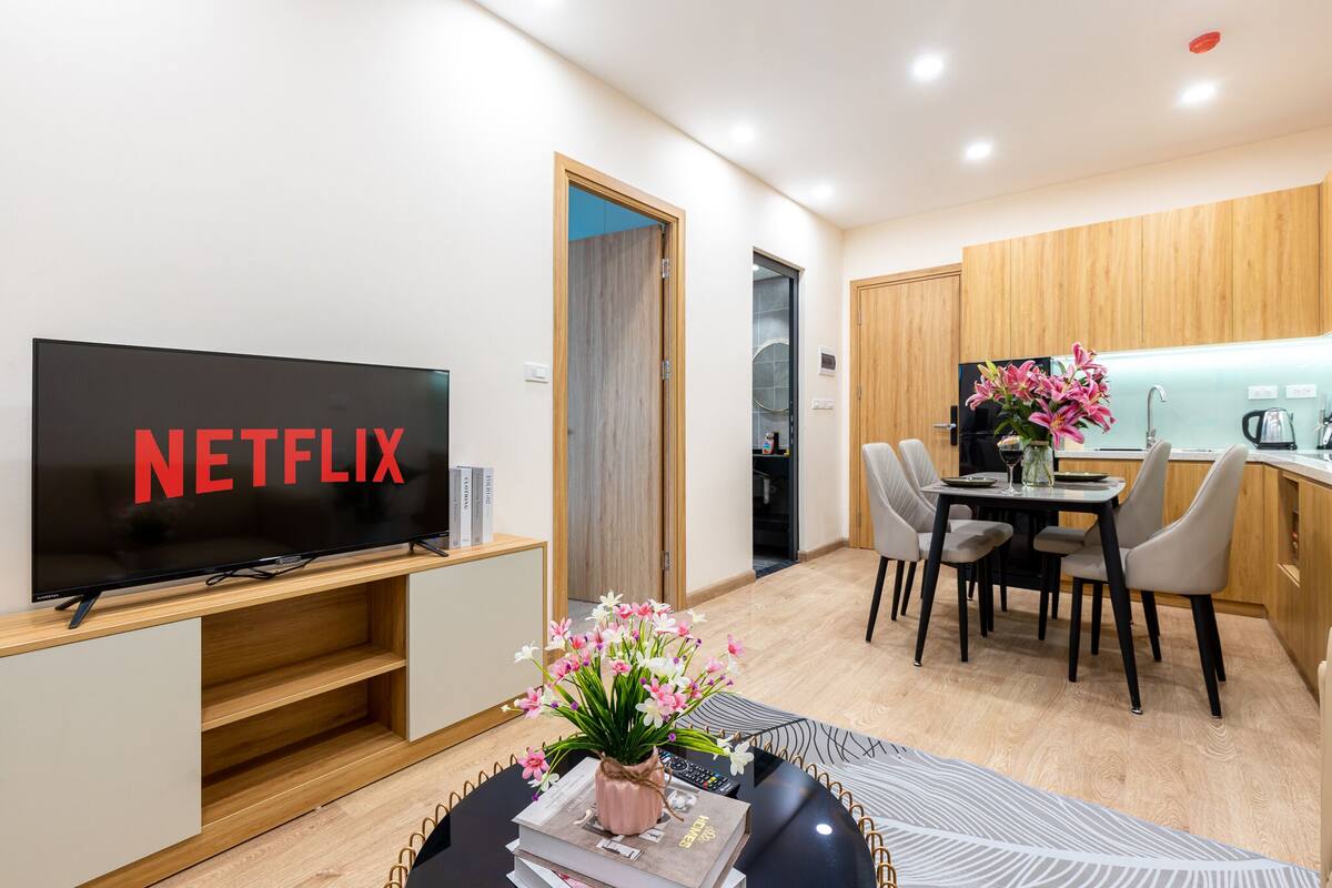 A modern living room with a flat-screen TV displaying "NETFLIX," a dining area with a table and chairs, and a kitchen with wooden cabinets and a sink.