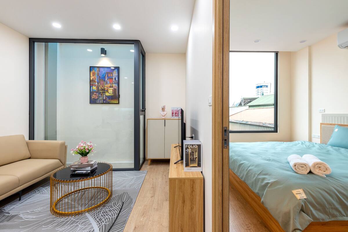 A modern hotel room with a sofa, a coffee table, a bed with rolled towels, a small cabinet, and a painting on the wall.