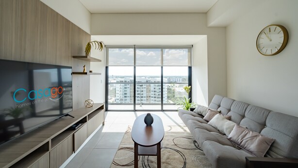 A modern living room with a large TV, a sofa, a coffee table, and a view of the city through large windows. - Tower Indico by Casago (Mérida)
