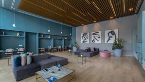 A modern lounge area with a sectional sofa, a coffee table, and a seating area with chairs. There are wall-mounted shelves and a large potted plant. - Tower Indico by Casago (Mérida)